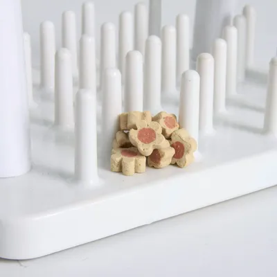 Gioco interattivo bianco con pioli verticali e croccantini a forma di fiore con centro rosso posizionati tra i pioli.