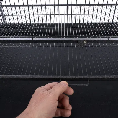 Hand opent een zwart metalen traliedeur met handgreep, onderdeel van een kooi of verblijf.