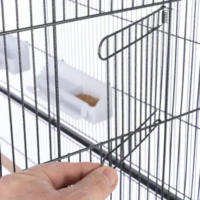 Close-up van een vogelkooi met metalen tralies en een hand die een deur opent. Op de achtergrond is een witte voerbak met zaden zichtbaar.