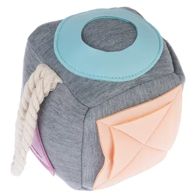 Cube en tissu gris avec éléments colorés, corde épaisse blanche sur un côté, cercle bleu et poche orange visibles. Aucun texte ou marque apparente.