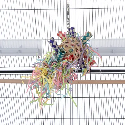 Jouet suspendu en osier avec bandes colorées et lamelles de papier multicolores, accroché à une cage par une chaîne métallique.