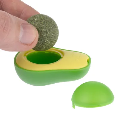 Jouet en forme d’avocat vert et jaune, une main tient une boule ronde verte à placer dans l’ouverture centrale du jouet. Coque en plastique démontée visible à côté.