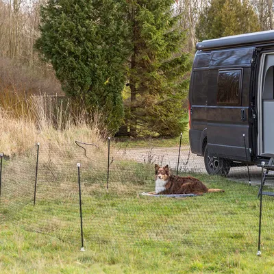 Mobiler Zaun aus Netzmaterial mit schwarzen Pfosten, aufgestellt neben einem Camper. Hund liegt innerhalb des umzäunten Bereichs auf einer Decke.