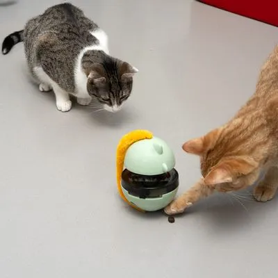 Dois gatos junto a brinquedo interativo dispensador de comida, com um gato a tocar numa guloseima caída no chão. Brinquedo em plástico verde claro e amarelo.