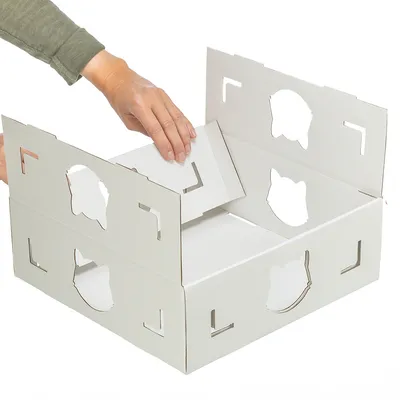 Hand zeigt Aufbau einer weißen Kartonbox mit Katzenkopf-Ausschnitten. Anleitung zur Montage des Produkts.