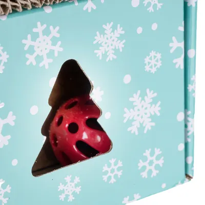 Rote Spielkugel sichtbar durch einen Tannenbaum-Ausschnitt in einer türkisfarbenen Verpackung mit Schneeflocken-Muster.
