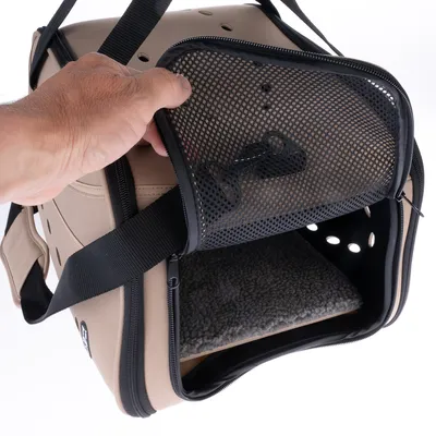 Sac de transport beige pour animaux avec ouverture en maille noire, poignée, sangle et coussin intérieur gris visible. Main tenant l’ouverture zippée.