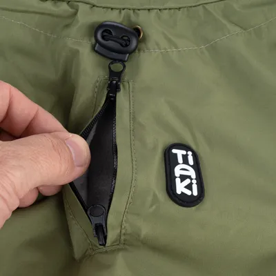 Poche zippée sur tissu vert avec badge Tiaki visible, main ouvrant la fermeture éclair. Cordon de serrage noir en haut de l’image.
