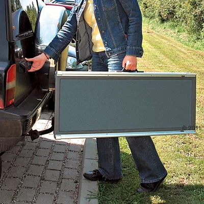 Osoba drží skládací rampu s kovovým rámem a tmavým povrchem u auta, připravena k použití nebo přepravě.