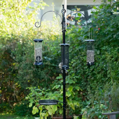 Vogelfutterstation mit drei hängenden Futterspendern für Samen und Körner, sowie einer Plattform für zusätzliche Fütterung. Metallgestell in einem Gartenbereich.