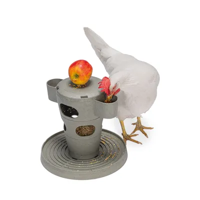 Comedero gris con varias aberturas, lleno de granos. Una gallina blanca picotea una abertura. Una manzana roja está encima del comedero.