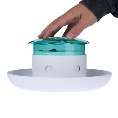 Hand drückt auf grünen Deckel eines weissen Futterspenders mit runder Basis. Demonstration der Bedienung des Spenders.