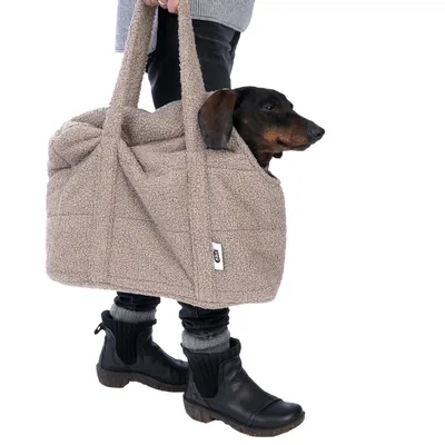 Sac de transport en tissu beige avec anses, marque visible Trixie, porté à la main, chien noir à l’intérieur, partie du corps humain visible jusqu’aux genoux.