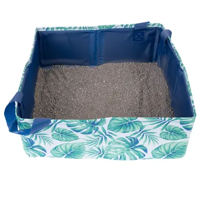 Transportowalna kuweta dla kota z tropikalnym wzorem liści, wypełniona żwirkiem. Boczne ścianki z niebieskiej tkaniny i uchwyty do przenoszenia.