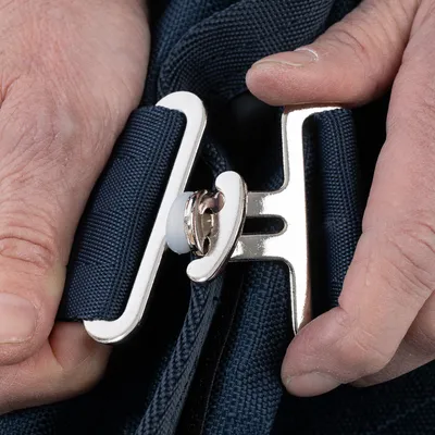Nahaufnahme eines silbernen Metallverschlusses an einem dunkelblauen Stoffband, gehalten von zwei Händen.