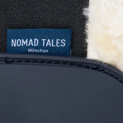 Étiquette bleue avec texte : NOMAD TALES München, cousue sur tissu noir à côté d’une surface beige duveteuse et d’un bord en matériau synthétique bleu foncé.