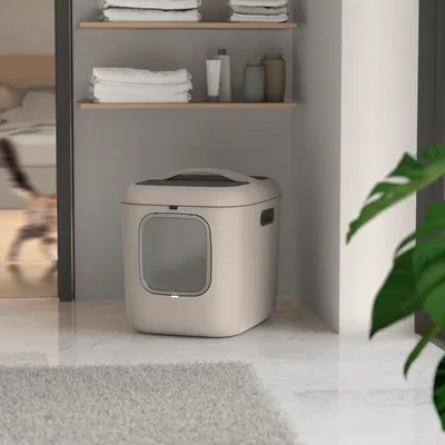 Beige Katzenklo mit Sichtfenster und abnehmbarem Deckel, platziert in einer modernen Wohnung neben Regalen mit Handtüchern und Dekorationsgegenständen.