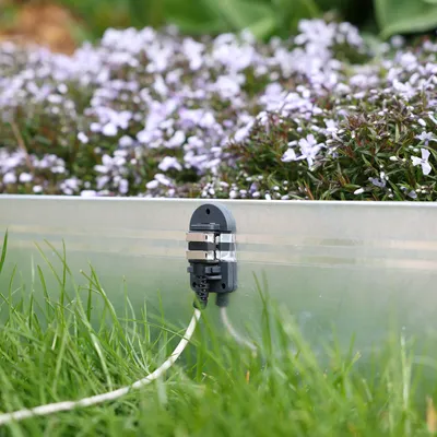 Elektrischer Anschluss an einem Metallrahmen, umgeben von Gras und Blumen. Kabel führt vom Anschluss weg.