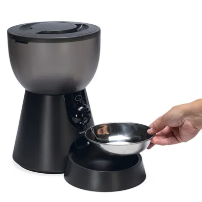Automatische voerdispenser met zwart design, uitneembare roestvrijstalen kom en bedieningsknop. Hand toont het uitnemen van de kom.