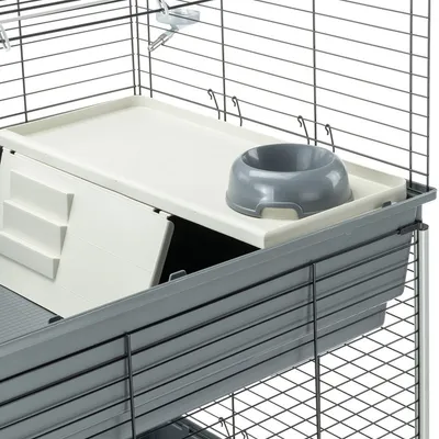 Käfig mit grauem Futternapf und weißer Plattform, darunter eine verschließbare Klappe. Metallgitterwände und Haken sichtbar.
