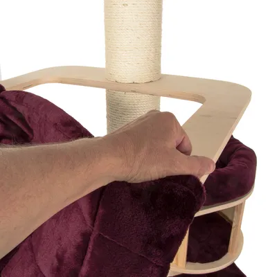 Hand demonstrating how to attach or remove a burgundy fabric cover from a wooden platform on a scratching post structure.