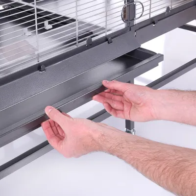 Persona mostrando la bandeja extraíble de una jaula metálica para facilitar la limpieza. Estructura y materiales visibles: metal.