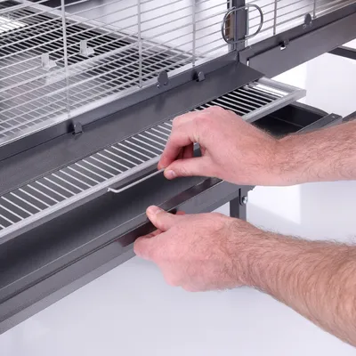 Manos extrayendo la bandeja inferior metálica de una jaula gris con rejilla, mostrando el sistema de limpieza extraíble.