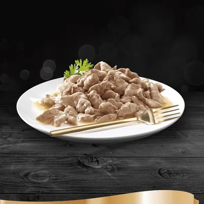 Prato branco com pedaços de comida húmida em molho, decorado com uma folha verde e acompanhado por um garfo dourado. Não são visíveis marcas ou texto.