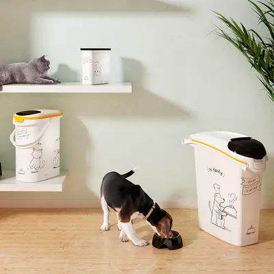 Contenedores de pienso blancos con dibujos de gatos y perros, uno grande y otro mediano. Un perro come de un cuenco negro y un gato gris está tumbado en una estantería.