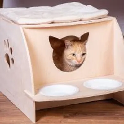 Cuccia in legno chiaro con ingresso a forma di testa di gatto, cuscino superiore e due ciotole bianche integrate sul davanti, decorazione laterale a zampa.
