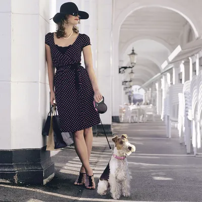 Donna con abito a pois e cappello tiene al guinzaglio un cane di taglia media con collare rosa e guinzaglio avvolgibile, borse per acquisti in mano, ambiente urbano elegante.