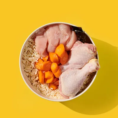 Bowl containing raw chicken pieces, a chicken drumstick, sliced carrots and white rice, showing visible ingredients used in the product.