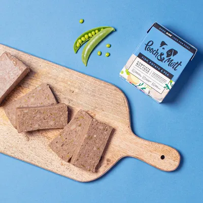 Sliced fish, potato and pea wet food shown on a wooden board next to packaging and fresh peas, illustrating product ingredients and texture.