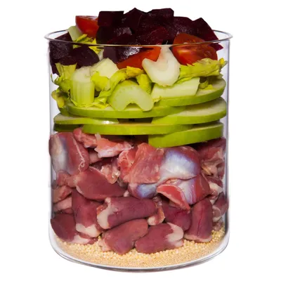 Glass container with visible layers of raw meat, green apple slices, celery, cherry tomatoes, beetroot pieces and small yellow grains at the bottom. All ingredients clearly separated.