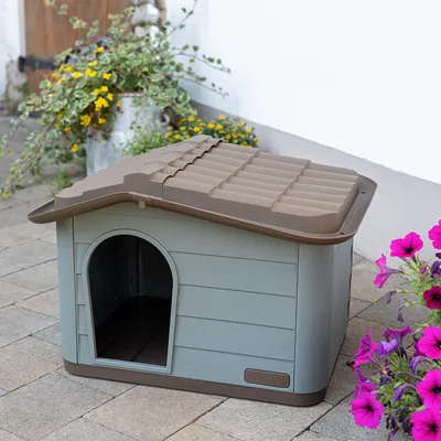 Graues Kunststoff-Hundehäuschen mit braunem Dach und ovalem Eingang, platziert auf Steinboden neben blühenden Blumen.