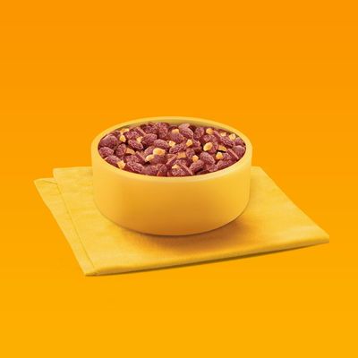 Bowl filled with dry pet food pieces and small chunks, placed on a folded yellow cloth against an orange background.