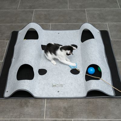 Play mat made of grey felt with holes, cat playing with blue toy and colourful balls. Modern Living brand visible on the mat.