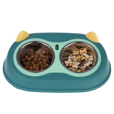 Gamelle double Tiaki avec deux bols en métal, l’un rempli de croquettes, l’autre de biscuits en forme d’os. Support en plastique vert avec oreilles jaunes visibles.