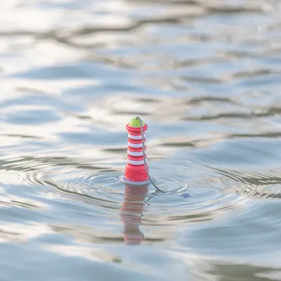 Jouet flottant en forme de phare rouge et blanc avec une corde, posé sur l’eau. Matériau plastique visible.