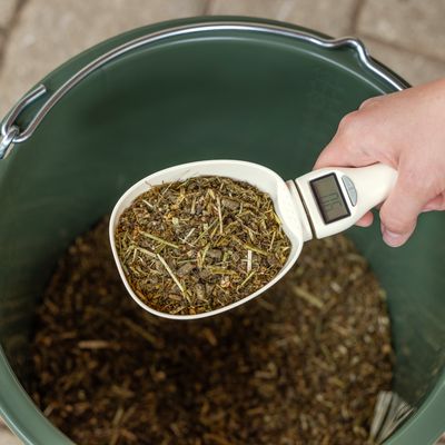 Hand håller ett foderskopa med inbyggd digitalvåg, fylld med hö och pellets över en grön hink.