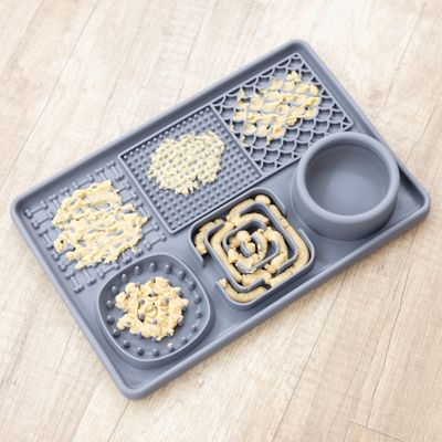 Graue Fütterungsmatte mit verschiedenen Texturen und Mustern, teilweise mit Futter bestrichen. Enthält auch eine runde Schale für Flüssigkeiten oder Snacks.