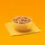 Bowl filled with small chunks of wet pet food in gravy, placed on a folded yellow cloth against an orange background.