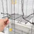 Main ouvrant une porte sur cage en métal noir avec perchoir, abreuvoir transparent à base jaune et mangeoire transparente fixés aux barreaux.