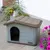 Graues Kunststoff-Hundehäuschen mit braunem Dach und ovalem Eingang, platziert auf Steinboden neben blühenden Blumen.