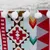 Detailní záběr na látkový design s geometrickými vzory v červené, hnědé, žluté a tyrkysové barvě, s bílými bambulkami a chlupatým okrajem.