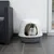 Chat blanc et noir assis dans une maison de toilette pour chat en plastique gris et blanc, forme arrondie, entrée large à l’avant. Mobilier d’intérieur visible en arrière-plan.