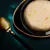Close-up of a bowl with light-coloured soup, visible pieces of chicken and vegetables. Next to it, a golden spoon on a dark background.