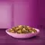 Bowl filled with mixed dry pet food pieces in various shapes and colours, including brown, yellow, green, and orange, displayed on a purple surface.