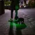 Petit chien avec collier et laisse lumineux verts, tenu par une personne sur un trottoir la nuit. Collier et laisse éclairés pour visibilité nocturne.