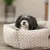 Petit chien noir et blanc allongé dans un panier pour chien en tissu beige à motif tressé, intérieur rembourré visible.